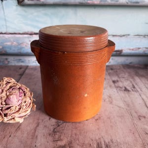 May include: A small, round, brown ceramic jar with a matching lid. The jar has two small handles and a textured surface. The lid sits snugly on top. The jar is likely vintage or antique, and is on a wooden surface.