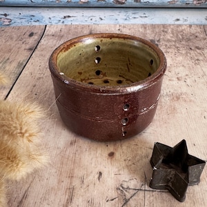 May include: A rustic, brown ceramic colander with multiple drainage holes. The interior has a greenish-yellow glaze. A star-shaped metal cookie cutter sits nearby on a wooden surface. The colander is a kitchenware item.