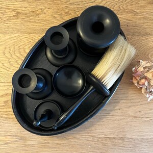 May include: A black wooden tray containing five black grooming tools, including a brush, a shaving bowl, and three small containers. The tray is on a wooden surface.