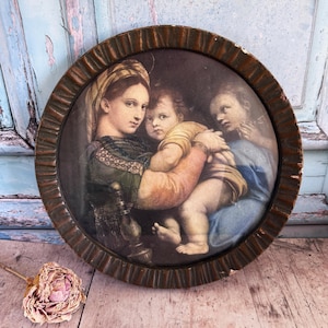 Puede incluir: Una impresión enmarcada redonda de una pintura de estilo renacentista. La obra de arte representa tres figuras, incluida una mujer que sostiene a un niño. El marco es marrón oscuro y texturizado. La pintura utiliza colores apagados.