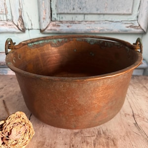 May include: A round copper pot with a wide opening and a curved handle on each side. The exterior surface shows a hammered texture with visible signs of oxidation and green patina along the upper rim and the metal handle attachments.