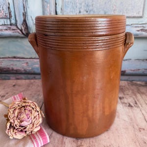 以下が含まれることがあります： 上部にリブのデザインと側面にハンドルが付いた、蓋付きの茶色の炻器の壺。アンティークのキッチン用品は、乾燥したピンクの花とストライプの布と一緒に木製の表面に展示されています。