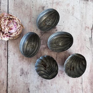 May include: Five antique metal moulds are displayed on a weathered wooden surface. The moulds are in oval and seashell shapes, with a dark, aged appearance. A dried, pink rose lies nearby. The moulds are likely for baking or decorative use.