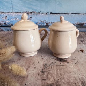 Puede incluir: Dos tarros de cerámica blanquecina con tapa y asas. Los tarros tienen forma redondeada con una base pequeña y una tapa con un pomo. El fondo es una superficie azul desgastada.