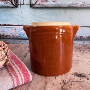 May include: A brown ceramic crock with a lid, featuring two side handles. The crock has a cylindrical shape with horizontal ridges near the top. The lid is a lighter tan color. The background includes a wooden surface and a distressed blue wall.