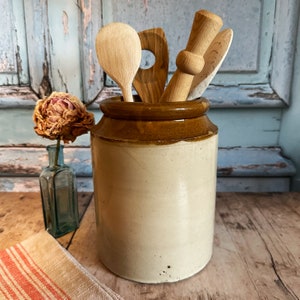 May include: A brown and white ceramic crock with a brown rim, filled with wooden kitchen utensils, including a wooden spoon, a wooden spatula, and a wooden stirring stick.