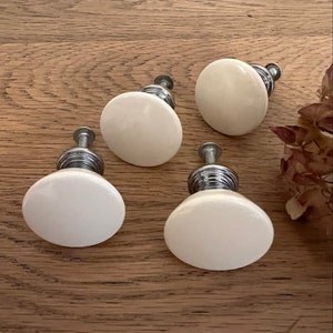 May include: Four white ceramic cabinet knobs with silver metal bases. The knobs are round and have a smooth, glossy finish.