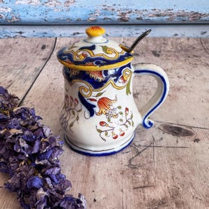 Puede incluir: Azucarero de cerámica blanca con tapa y cucharita. El azucarero está decorado con motivos florales azules, amarillos y rojos. El asa es blanca con ribetes azules. El azucarero mide unos 10 cm de alto.