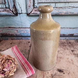 Puede incluir: Una botella de gres vintage con una parte superior redondeada y un cuerpo cilíndrico. La botella es de color marrón claro con una banda más oscura en el medio. Un paño a rayas rosas y blancas y una flor seca están en primer plano.