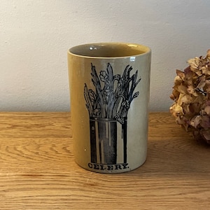 Może przedstawiać: Gliniany słoik w kolorze beżowym z czarno-białym rysunkiem łodyg selera w puszce. Słoik ma napis "CELERY" nadrukowany na spodzie.