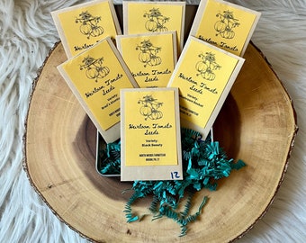 May include: A set of 12 heirloom tomato seed packets in a small box. The packets are yellow with black text and illustrations of tomatoes. The text on the packets reads "Heirloom Tomato Seeds" and lists the variety of tomato seeds inside. The packets are arranged in a box with green shredded paper.