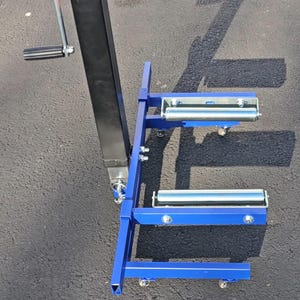 Puede incluir: Soporte de metal azul con cuatro ruedas y dos rodillos. El soporte tiene un poste de metal negro con un asa.