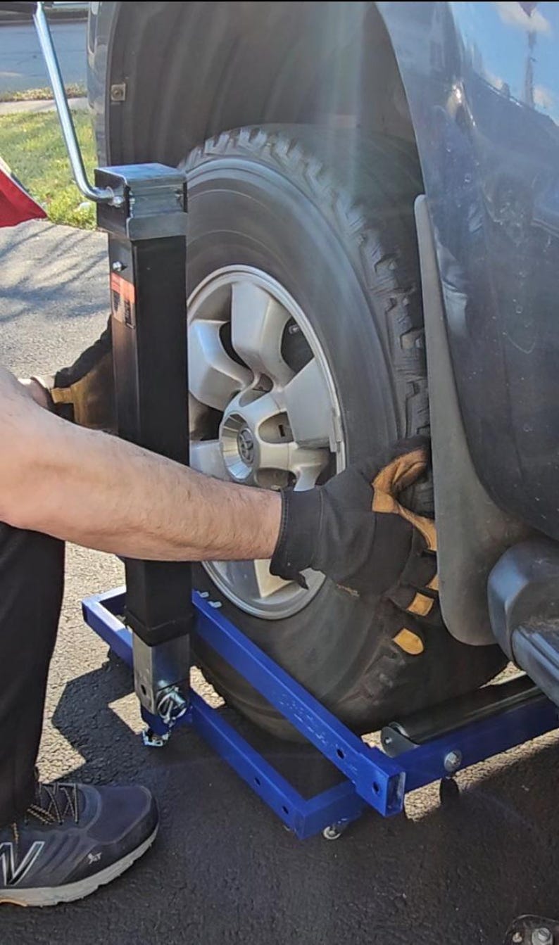 Puede incluir: Un gato de coche de metal azul con un gato hidr&aacute;ulico negro y una rueda encima. El gato tiene dos ruedas a cada lado y una manija para subir y bajar el gato.