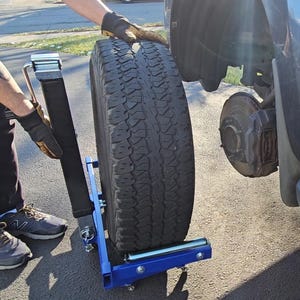 Puede incluir: Un carro de neum&aacute;ticos de metal azul con un neum&aacute;tico negro en &eacute;l. El carro tiene un asa y dos ruedas. Una persona est&aacute; usando el carro para mover el neum&aacute;tico.
