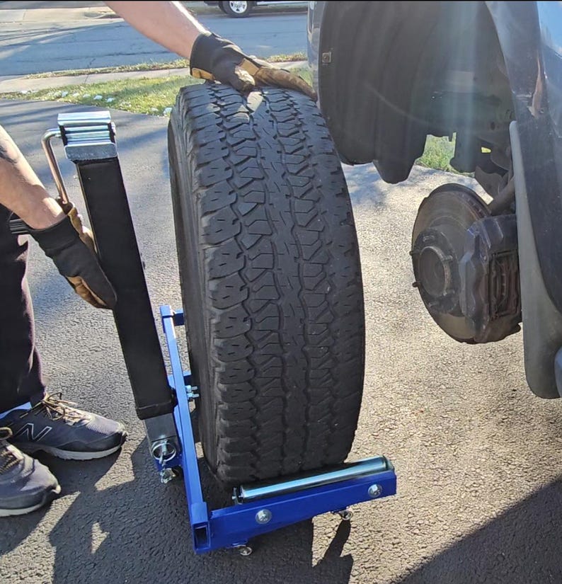 Puede incluir: Un carro de neum&aacute;ticos de metal azul con un neum&aacute;tico negro en &eacute;l. El carro tiene un asa y dos ruedas. Una persona est&aacute; usando el carro para mover el neum&aacute;tico.