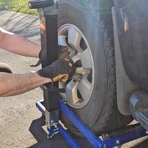 Puede incluir: Una persona usando un gato de cambio de neum&aacute;ticos azul y negro para quitar un neum&aacute;tico de un SUV negro. El gato tiene un mango de metal y una base de metal con ruedas.