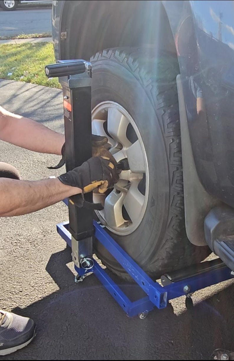 Puede incluir: Una persona usando un gato de cambio de neum&aacute;ticos azul y negro para quitar un neum&aacute;tico de un SUV negro. El gato tiene un mango de metal y una base de metal con ruedas.