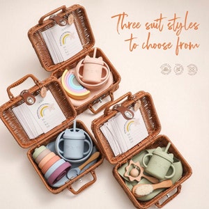 Puede incluir: Tres cestas de mimbre con tapas, cada una con un juego de alimentación para bebés diferente. El primer juego incluye un juguete de apilar arcoíris, un tazón de silicona rosa y una taza de silicona rosa con una pajita. El segundo juego incluye un tazón de silicona azul, una taza de silicona azul con una pajita, un juego de tazones de silicona apilables y una cuchara de madera. El tercer juego incluye una taza de silicona verde con una pajita, un sonajero de madera, un cepillo de madera y un plato de silicona verde.