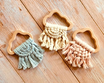 May include: Three wooden cloud-shaped teethers with macrame fringe. The teethers are in natural wood with a light blue, cream, and pink macrame fringe.
