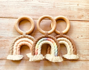 May include: Three macrame rainbow decorations with wooden rings. The rainbows feature a variety of colors, including beige, pink, and brown, with a fringe at the bottom. The wooden rings are attached to the top of each rainbow.