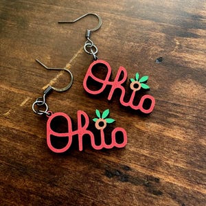 May include: A pair of red earrings featuring the word Ohio in a cursive font with a small orange flower and green leaves attached to the letter O. The earrings hang from dark metal hooks and are designed for pierced ears with a dangling style.