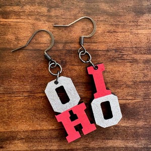 May include: A pair of dangle earrings featuring the letters O, H, I, and O. The letters are arranged in two pairs, with one earring displaying a silver O and red H, and the other displaying a red I and silver O. Each letter has a block font style.