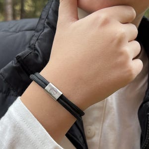 May include: A black rope bracelet with a silver clasp featuring a basketball design. The bracelet is worn on a wrist, with a black vest and white shirt in the background. The bracelet appears to be approximately 1.3 cm wide.