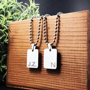 May include: Two silver necklaces with rectangular pendants. The pendants are engraved with the initials "J.Z." and "N". The necklaces are displayed on a wooden surface.