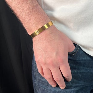 May include: A gold metal bracelet with a small cross design. The bracelet is worn on the left wrist.