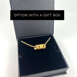 Peut inclure: Un collier en or avec une cha&icirc;ne et cinq breloques rondes dans une bo&icirc;te cadeau noire. La bo&icirc;te porte l'inscription "OPTION WITH A GIFT BOX" sur le dessus.