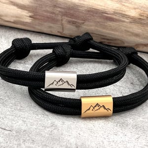 May include: Two black rope bracelets with mountain range designs. One bracelet has a silver-colored metal accent, and the other has a gold-colored metal accent. The bracelets are displayed on a light gray surface.