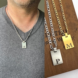 Peut inclure: Collier chaîne argenté avec un pendentif rectangulaire gravé de la lettre "P". Le collier est porté par une personne portant un pull gris. Un autre collier avec un pendentif doré avec la lettre "M" est également montré.