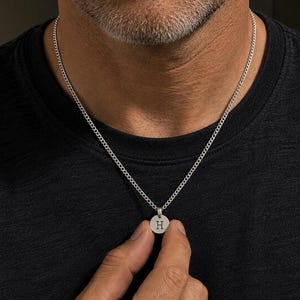 May include: A silver-toned necklace featuring a small, circular pendant engraved with the capital letter H. The pendant hangs from a delicate, fine-linked chain that rests against a dark-coloured shirt, showcasing a simple and clean design.