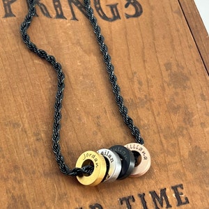Peut inclure: Un collier en cha&icirc;ne de corde noire avec quatre breloques rondes en m&eacute;tal. Les breloques sont dor&eacute;es, argent&eacute;es, noires et or rose. Chaque breloque porte un nom grav&eacute; : Jordan, Ailan, Lillian et Lilliana.