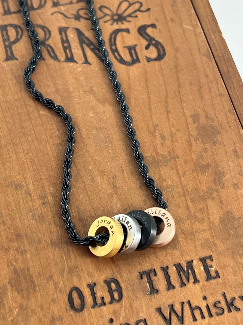 Peut inclure: Un collier en cha&icirc;ne de corde noire avec quatre breloques rondes en m&eacute;tal. Les breloques sont dor&eacute;es, argent&eacute;es, noires et or rose. Chaque breloque porte un nom grav&eacute; : Jordan, Ailan, Lillian et Lilliana.