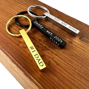 May include: Three gold, silver and black keychains with engraved bars. The gold keychain says "#1 DAD", the silver keychain says "YOU ARE HOME" with a heart symbol, and the black keychain says "Daddy est. 2024 Riley".