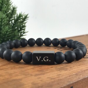 May include: Black beaded bracelet with a black metal bar engraved with the initials "V.G."