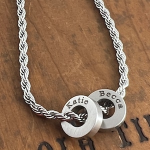 Peut inclure: Un collier en cha&icirc;ne de corde argent&eacute;e avec deux anneaux argent&eacute;s. Les anneaux sont grav&eacute;s avec les noms "Katie" et "Becca".