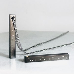 Peut inclure: Un collier en chaîne argentée avec deux pendentifs en forme de barre noire gravés de points braille. Les pendentifs sont rectangulaires et ont une finition polie.