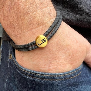 May include: Black rope bracelet with a gold-coloured clasp. The clasp features the letters "LG" in a black, minimalist font. The bracelet is worn on a wrist, with a dark denim pocket visible in the background.