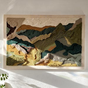 May include: A rectangular wall hanging featuring a textured punch needle landscape design in shades of forest green, olive, mustard yellow, beige, and brown. The artwork is mounted in a light-colored wooden frame and displays a mountain range.