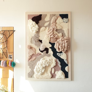 Könnte beinhalten: Ein gerahmter Wandbehang mit einem strukturierten abstrakten Design in Beige-, Rosa-, Weiß- und Dunkelblautönen. Das Design verfügt über erhabene, flauschige Formen und Texturen.