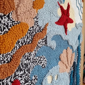 May include: A colorful abstract wall hanging made with yarn and a punch needle technique. The design features a variety of shapes and colors, including blue, red, orange, and white.
