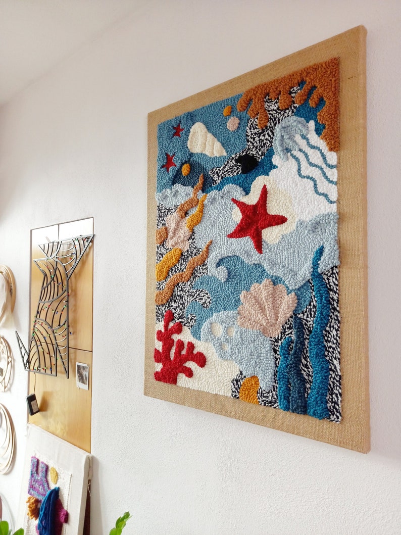 May include: A colorful wall hanging with a blue, white, and orange abstract design. The design features a starfish, seashells, and coral. The wall hanging is framed in a natural burlap fabric.