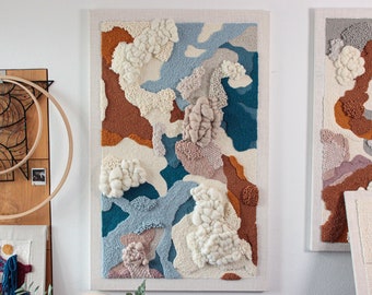 May include: A rectangular wall hanging featuring a textured punch needle design with abstract shapes in shades of cream, rust orange, light blue, and deep teal. The piece incorporates varying pile heights and tufted wool sections on a fabric base.