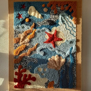 May include: A colorful abstract wall hanging with a blue background and various shapes and textures, including seashells, starfish, and coral. The wall hanging is made of yarn and is framed with a burlap border.