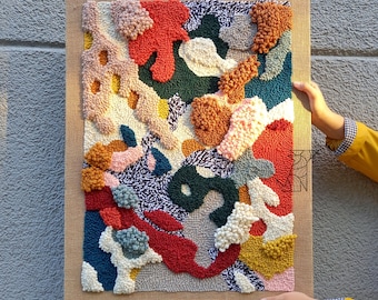 May include: A colorful abstract wall hanging made with yarn and a variety of textures. The piece features a mix of red, orange, yellow, green, blue, and white colors. The wall hanging is framed in a natural burlap material.
