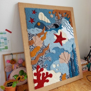 May include: A framed abstract wall art piece with a blue, white, and orange color scheme. The art is made of yarn and features a variety of shapes, including stars, seashells, and waves.