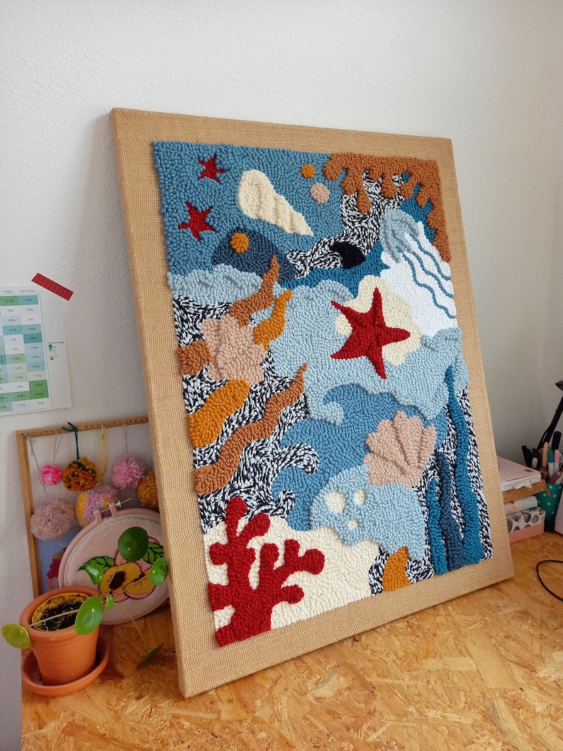 May include: A framed abstract wall art piece with a blue, white, and orange color scheme. The art is made of yarn and features a variety of shapes, including stars, seashells, and waves.