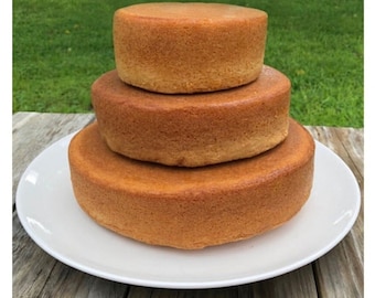 May include: Three round cake layers stacked on a white plate. The layers are a light brown color and have a slightly rough texture.